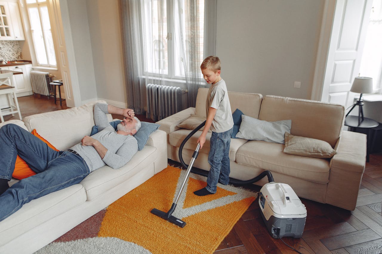 portfolio-01 A young boy vacuums the living room carpet while his grandfather relaxes on the sofa.